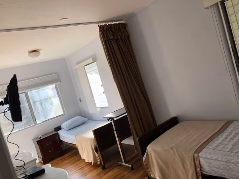 Two single beds in a bright room with wood floors, a privacy curtain, a bedside table, and a wall-mounted TV.