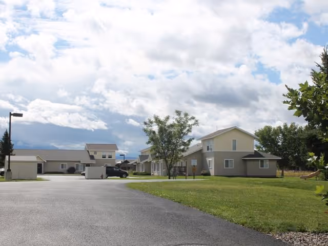 Exterior view of Rendezvous Apartments with lawns, drive, and parked cars along a street.