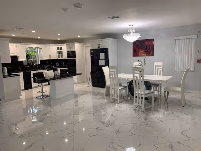 Open-plan bright white kitchen and dining area with a central island, marble-look tile floor, a white dining table and chairs, black appliances, and a chandelier.