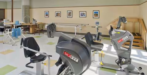 Bright rehab gym with exercise machines, chairs, and framed wall art.