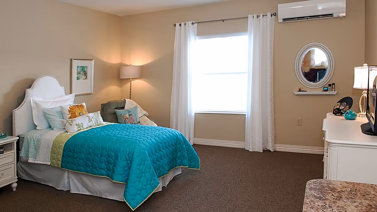A cozy bedroom with a bed featuring a white headboard, white and patterned pillows, and a teal quilt. There is a nightstand with a lamp and a small chair with cushions in the corner. A window with white curtains lets in natural light. Across from the bed is a white dresser with a round mirror above it and a TV on top. The walls are beige and the carpet is brown.