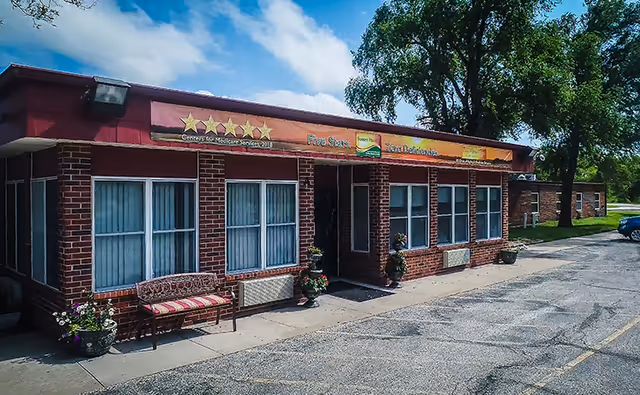 Exterior view of a single-story brick building with large windows covered by white curtains. There is a bench with a cushion and flower pots near the entrance. The building has a sign above the entrance displaying five stars and text indicating a five-star rating from the Centers for Medicare Services in 2018. Trees and a parking area are visible around the building under a partly cloudy sky.