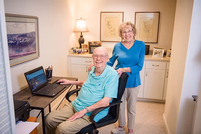 An elderly man sitting in an office chair at a desk with a laptop, smiling at the camera. An elderly woman stands behind him with her hand on his shoulder, also smiling. The room has a lamp, framed pictures on a cabinet, and artwork on the walls.