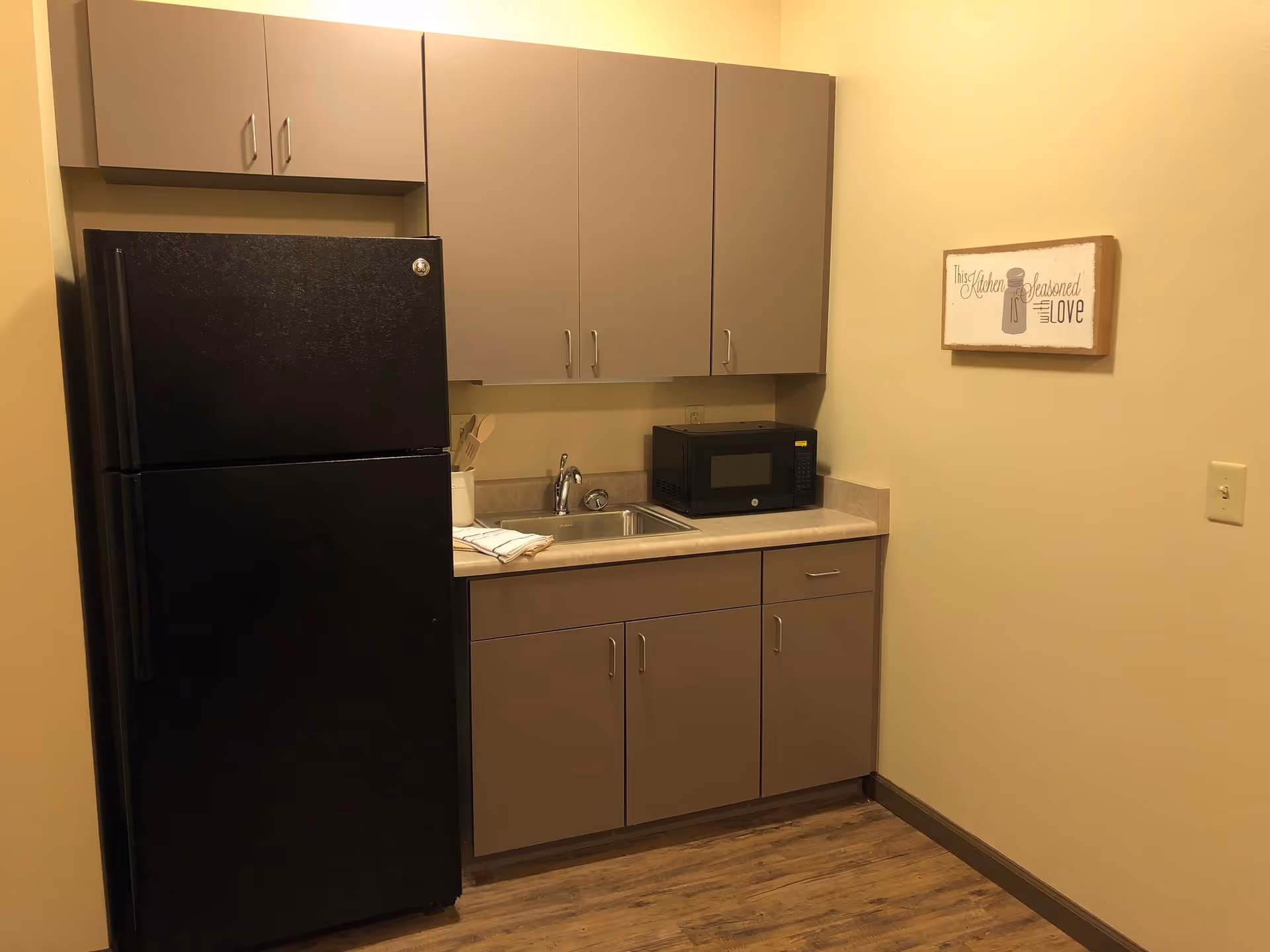 Small kitchenette with a black refrigerator, gray cabinets, a sink, microwave on the counter, and a framed wall sign.