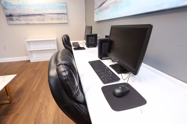 A modern computer workstation with two black leather chairs, two desktop monitors, keyboards, and mice on a white marble-patterned desk in a room with wooden flooring and light gray walls. There are two large abstract paintings on the walls and a small white shelf in the corner.