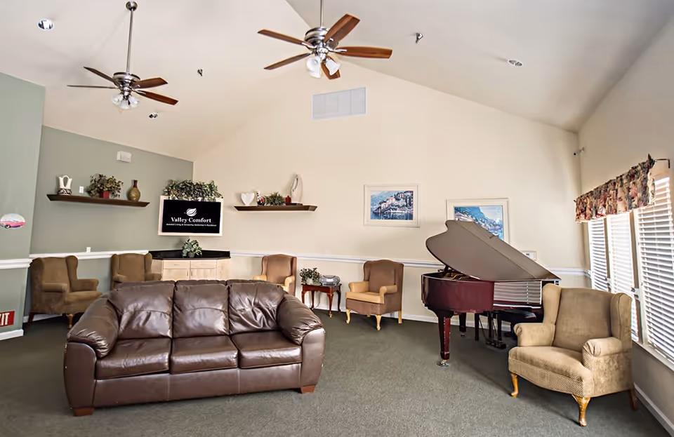 A spacious living room with a brown leather sofa in the center, surrounded by several upholstered armchairs. There is a grand piano near the windows with floral valances, two ceiling fans, and wall shelves with decorative items. A TV mounted on the wall displays the Valley Comfort logo.