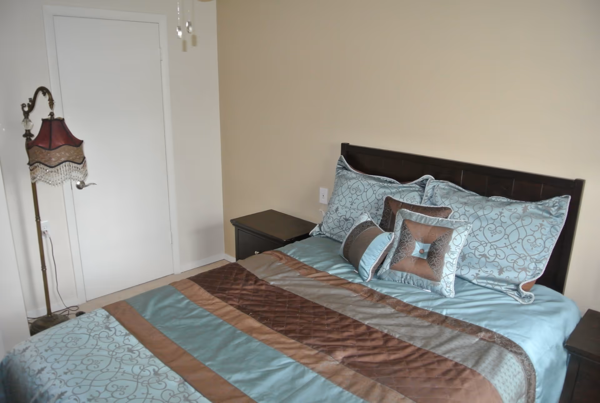 A bedroom with a bed made up with a blue and brown patterned comforter and matching pillows. There is a dark wooden headboard and two dark wooden nightstands on either side of the bed. A vintage-style floor lamp with a red and beige lampshade stands next to a closed white door.