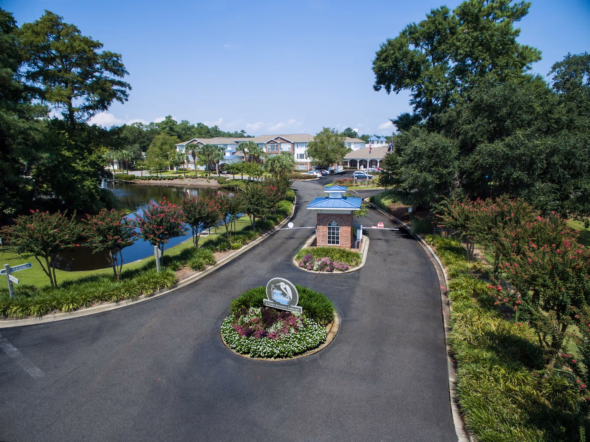 Entrance driveway with a small guardhouse and landscaped roundabout by a pond, leading to residential buildings surrounded by trees.