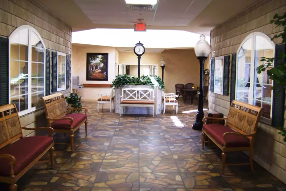 Indoor lobby hallway with benches, decorative clock, lamp posts, plants, and tiled floor in a senior living facility.