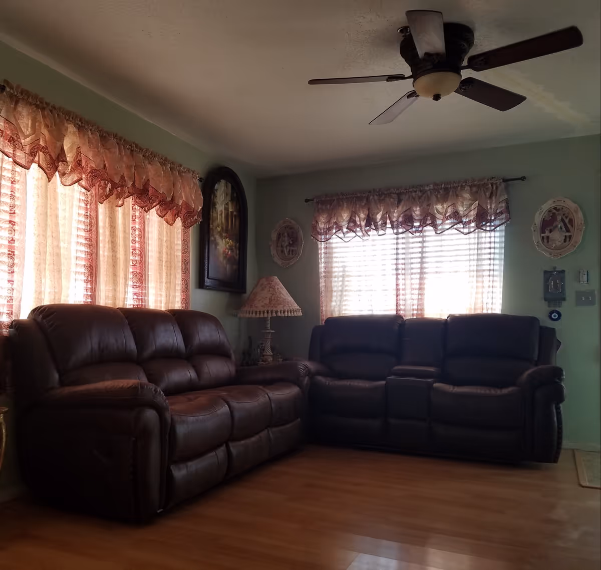 A living room with two dark brown leather sofas arranged in an L shape, a wooden floor, two windows with sheer curtains and valances, a ceiling fan with lights, a table lamp with a decorative shade, and framed wall art and decorations.
