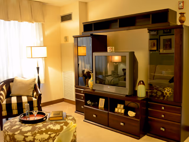 Cozy living room with a striped armchair, patterned ottoman, floor lamp, and a dark wood entertainment center holding a TV and decorative items.