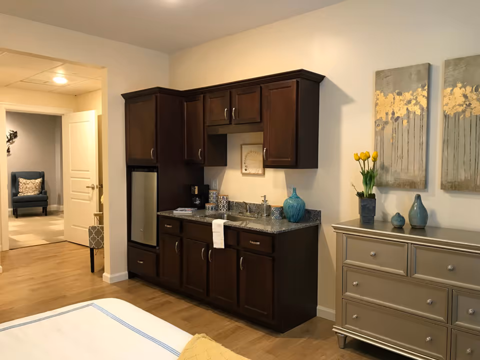 Interior view of a room in The Chelsea at Shrewsbury featuring a kitchenette with dark wood cabinets, a small sink, a mini refrigerator, and a coffee maker. To the right, there is a silver dresser with decorative vases and yellow tulips on top, and two abstract paintings with gold accents hanging on the wall. In the background, an open door leads to another room with a blue armchair and a decorative pillow.