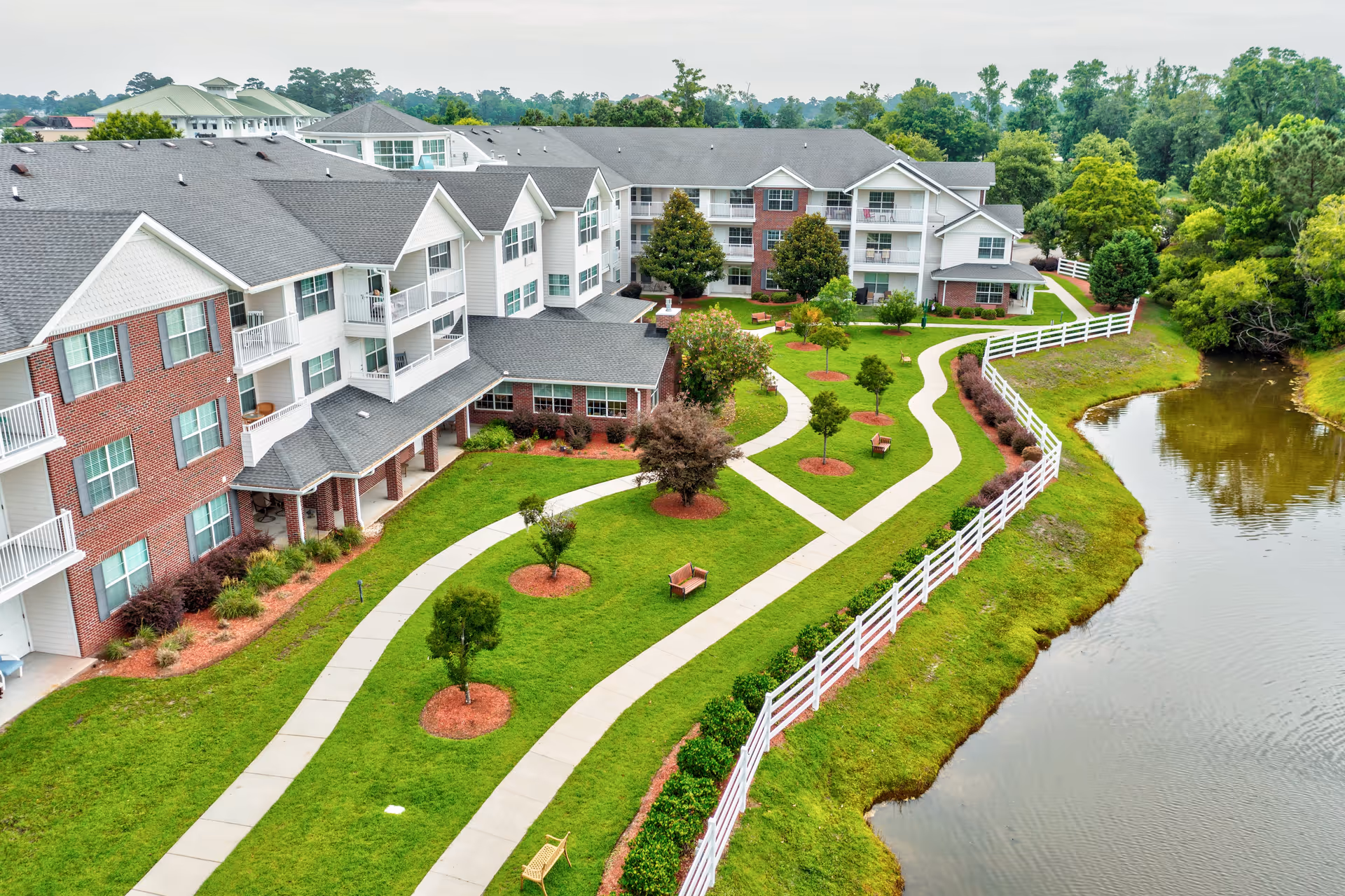 Aerial view of TerraBella Myrtle Beach senior living facility showing a large multi-story building with white and brick exterior. The building is surrounded by well-maintained green lawns, landscaped trees, walking paths, benches, white fencing, and a pond on the right side.