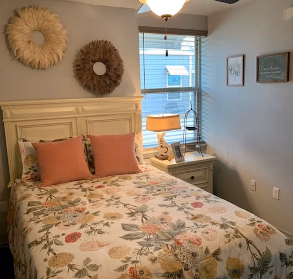 A cozy bedroom with a floral-patterned bedspread and two coral-colored pillows on a cream-colored wooden bed frame. There are two decorative round wall hangings above the bed, a window with blinds, a nightstand with a lamp, a photo frame, and a small decorative stand. Two framed pictures hang on the adjacent wall.