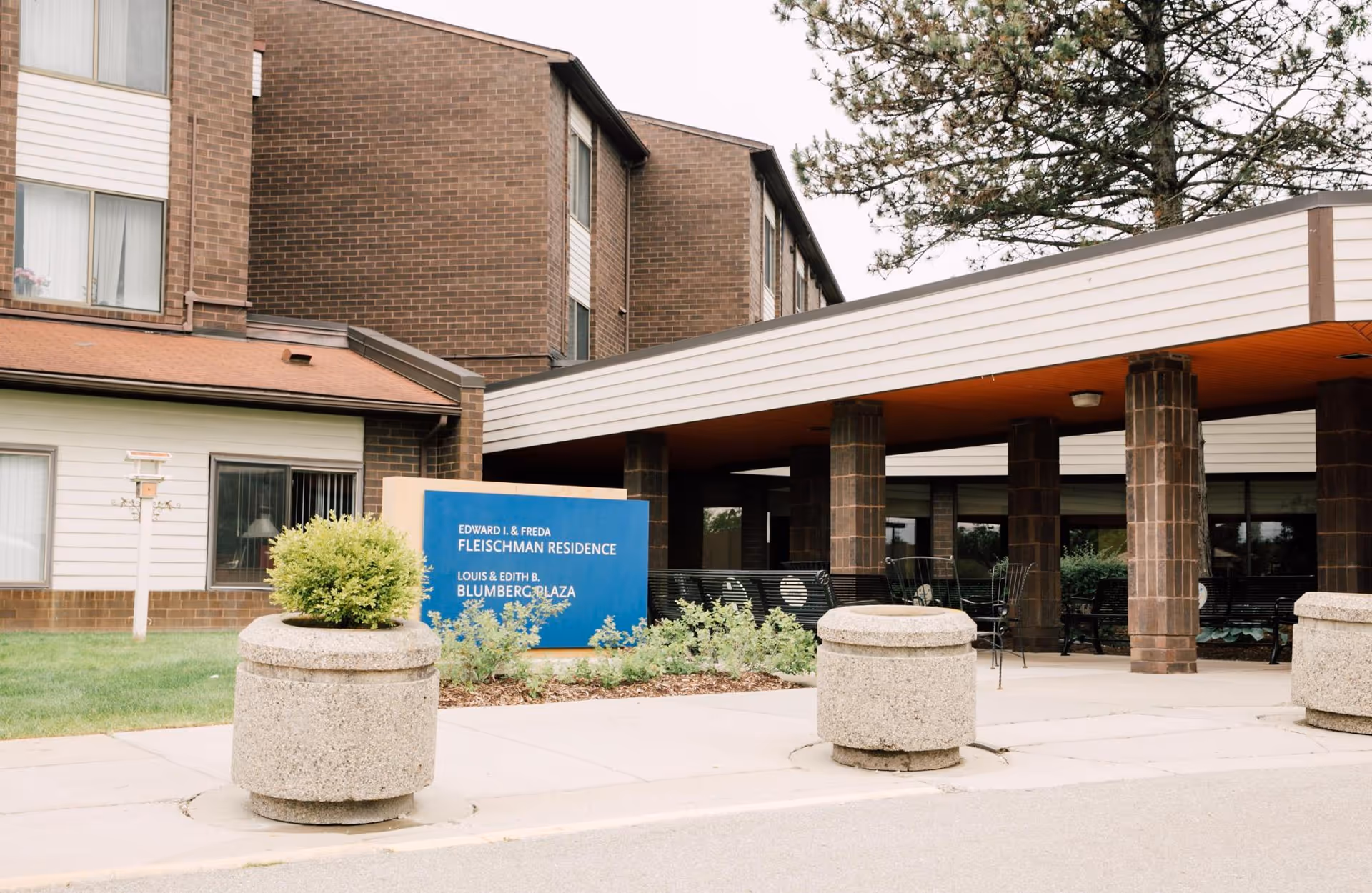 Exterior view of a senior living facility building with a covered entrance supported by brick columns. There is a blue sign in front of the entrance that reads 'Edward I. & Freda Fleischman Residence, Louis & Edith B. Blumberg Plaza'. Concrete planters and some greenery are visible near the sidewalk.