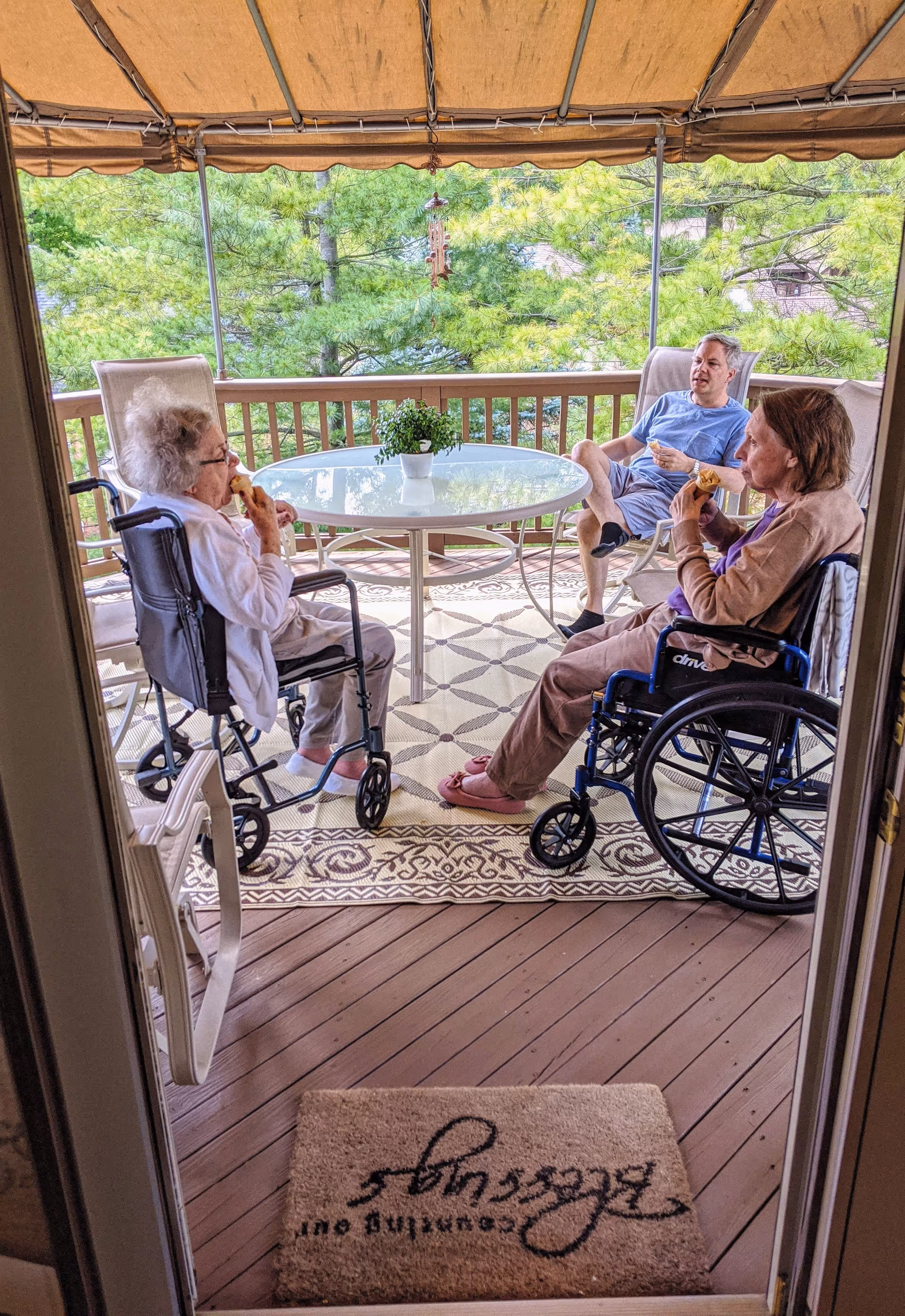 Two elderly women in wheelchairs and a man sitting on a covered outdoor balcony, enjoying snacks around a glass table with a potted plant, surrounded by trees and greenery.