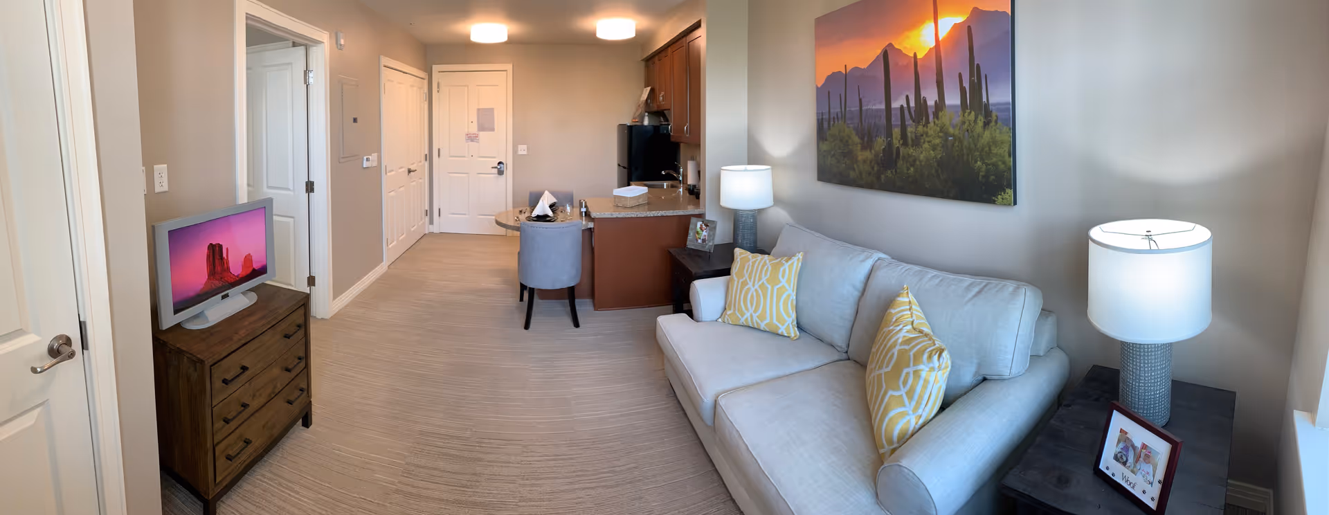 A cozy living area in a senior living facility with a light gray sofa adorned with yellow patterned pillows, a side table with a lamp and framed photo, a large wall art of a desert sunset with cacti, a small dining table with two chairs, and a kitchenette with wooden cabinets and a black refrigerator. A TV on a wooden dresser is visible near the entrance door.