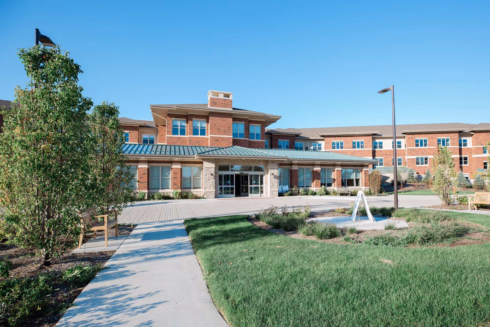 Exterior view of a senior living facility building with a brick facade, large windows, and a green metal roof. The entrance is centered with glass doors, surrounded by landscaped greenery, a paved walkway, and benches under a clear blue sky.