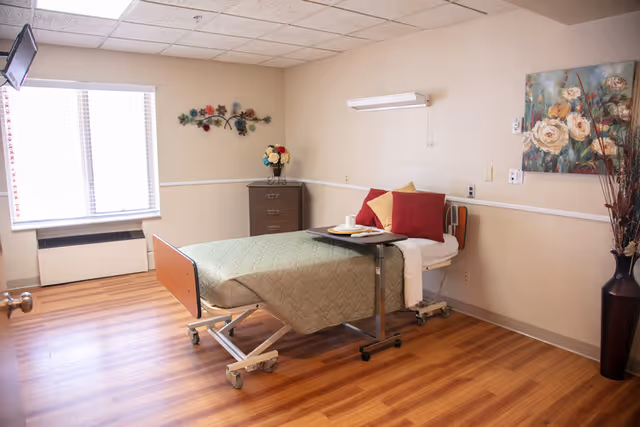 A simple, clean senior living bedroom with a single hospital-style bed covered with a green quilt and red and beige pillows. A small overbed table holds a cup and plate. The room has wood flooring, a window with blinds, a small chest of drawers with a flower arrangement, wall decorations including a floral painting and metal wall art, and a tall vase with decorative branches.
