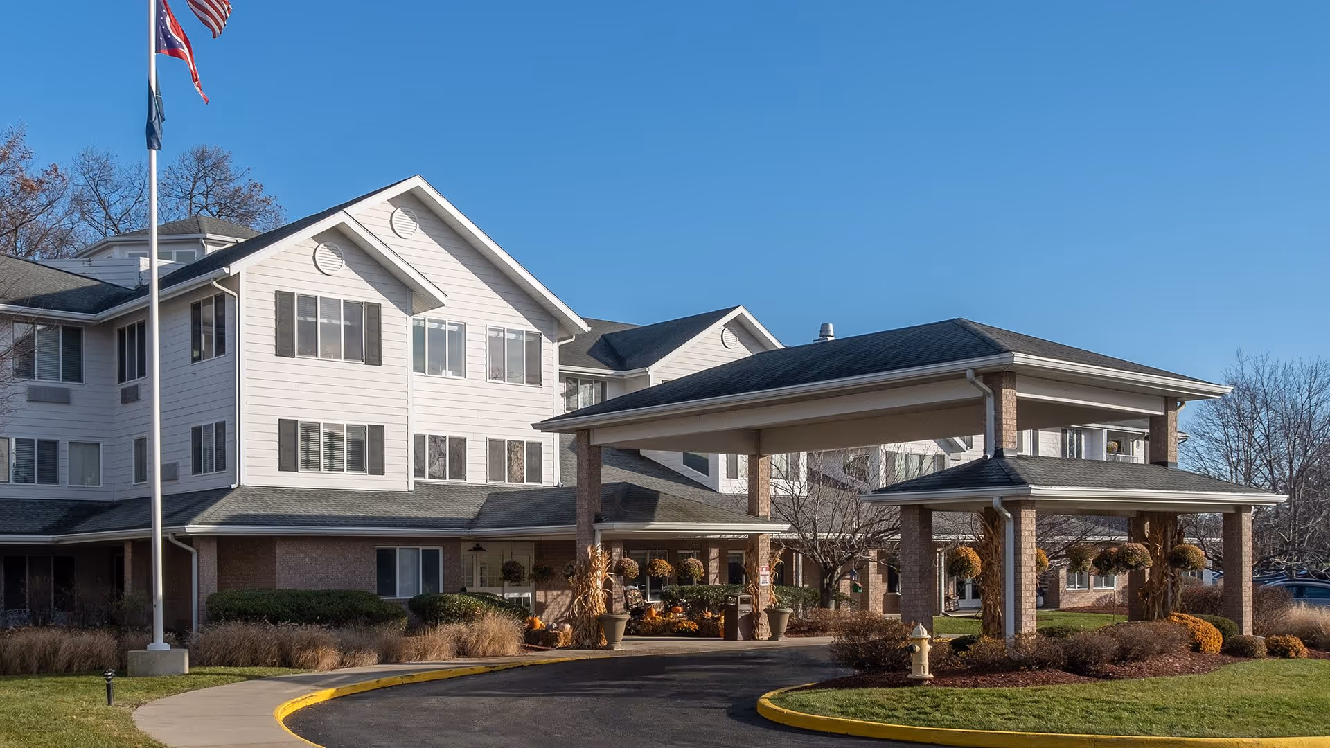 Exterior view of Holiday Alexis Gardens senior living facility showing a multi-story building with white siding and brick accents, a covered entrance with columns, landscaped bushes and grass, and an American flag on a flagpole against a clear blue sky.
