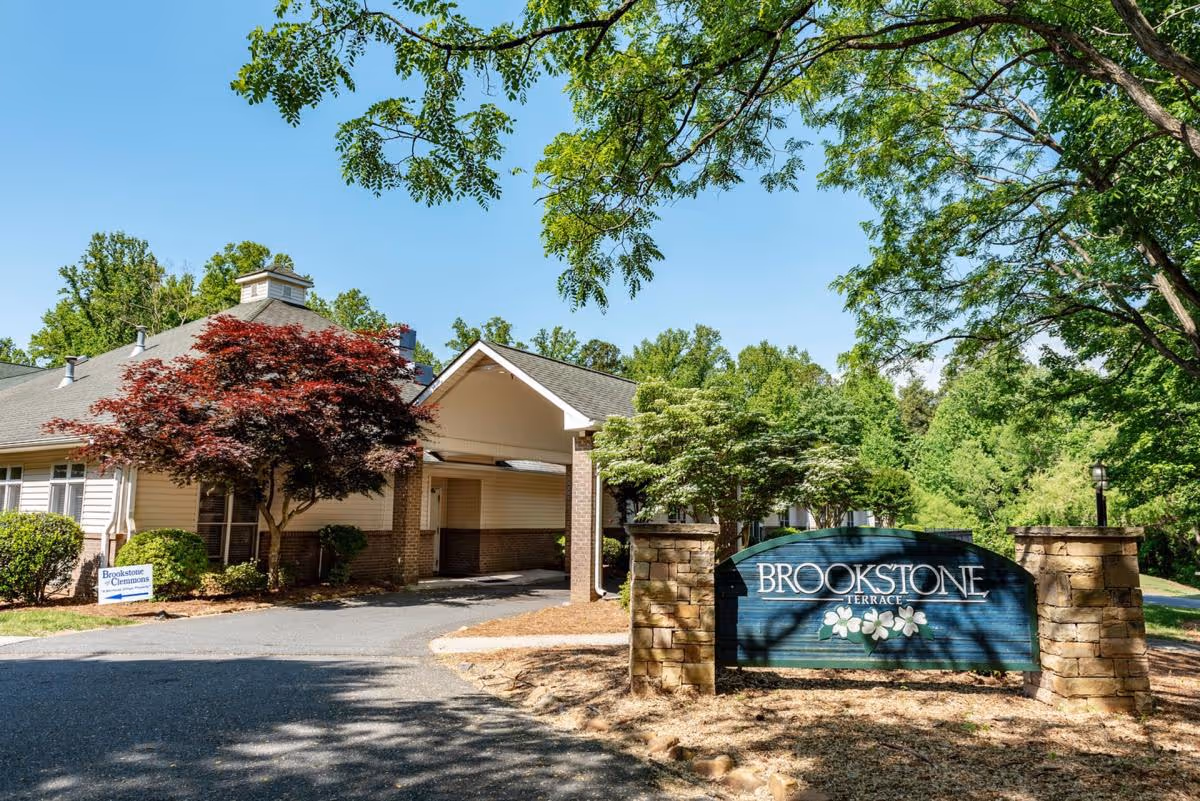 Exterior view of Brookstone Terrace senior living facility with a driveway, trees, and a large sign displaying the facility name surrounded by stone pillars.