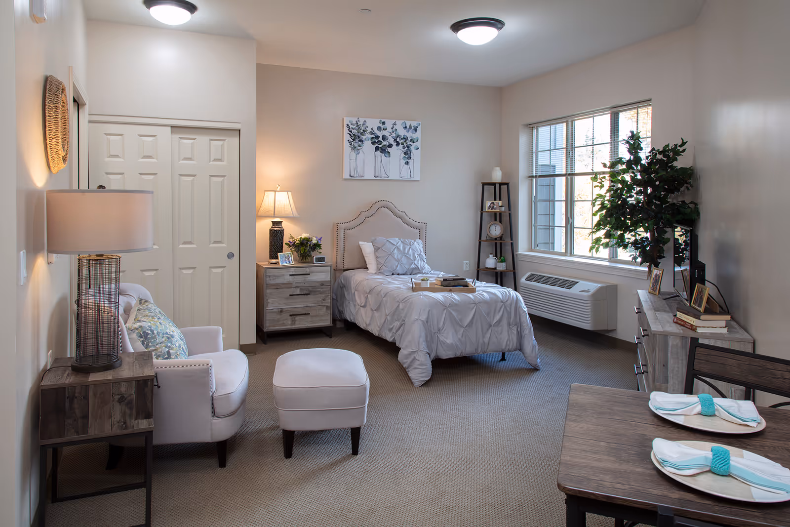 A cozy senior living bedroom with a single bed covered in a light gray comforter, a wooden nightstand with a lamp and flowers, a comfortable armchair with an ottoman, a wooden dresser with a TV and framed photos, a small dining table set for two, and a large window letting in natural light.