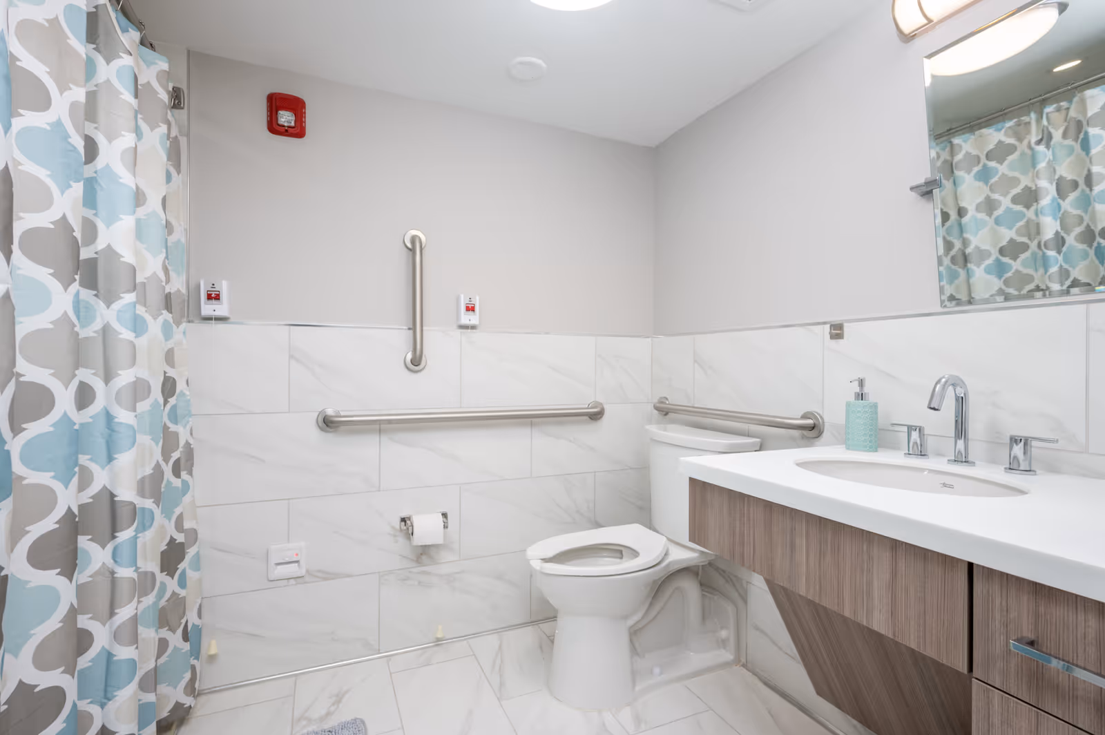 A clean and modern bathroom with white marble tiles on the walls and floor. The bathroom features a toilet with grab bars on the walls for accessibility, a sink with a large mirror above it, and a shower area with a patterned curtain in shades of blue, gray, and white. There are emergency pull cords on the wall near the toilet and shower.