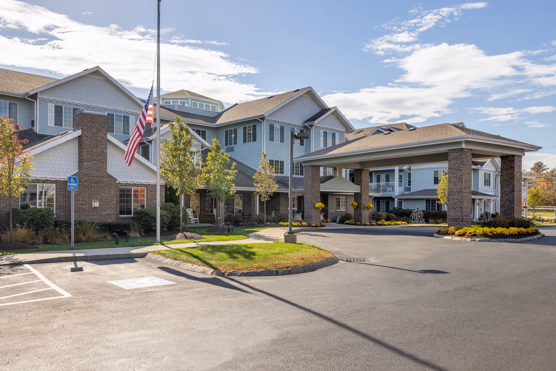 Exterior view of Holiday Kittery Estates senior living facility showing a large building with multiple windows, a covered entrance supported by brick columns, an American flag at half-mast, landscaped greenery, and a clear blue sky with some clouds.