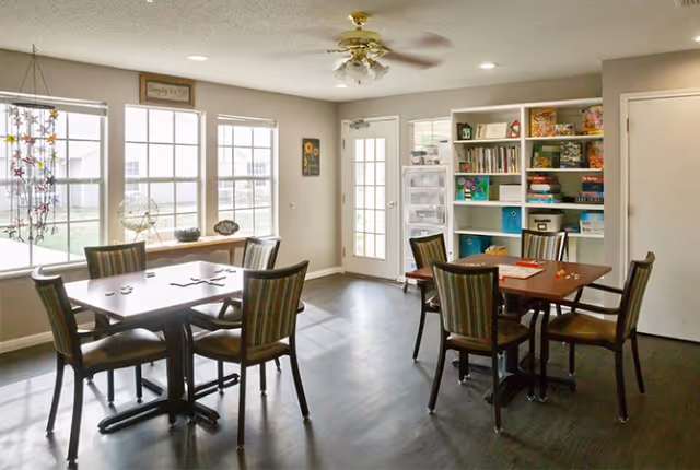 A bright room with two tables and several chairs arranged around them. The tables have board games and puzzles on them. There is a large window letting in natural light, a door with glass panes, and a white shelving unit filled with books and games. The floor is dark wood, and the walls are light-colored.