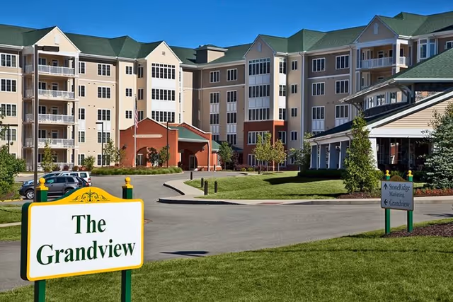 Exterior view of a multi-story senior living facility named The Grandview, with a driveway, green lawns, and clear blue sky.