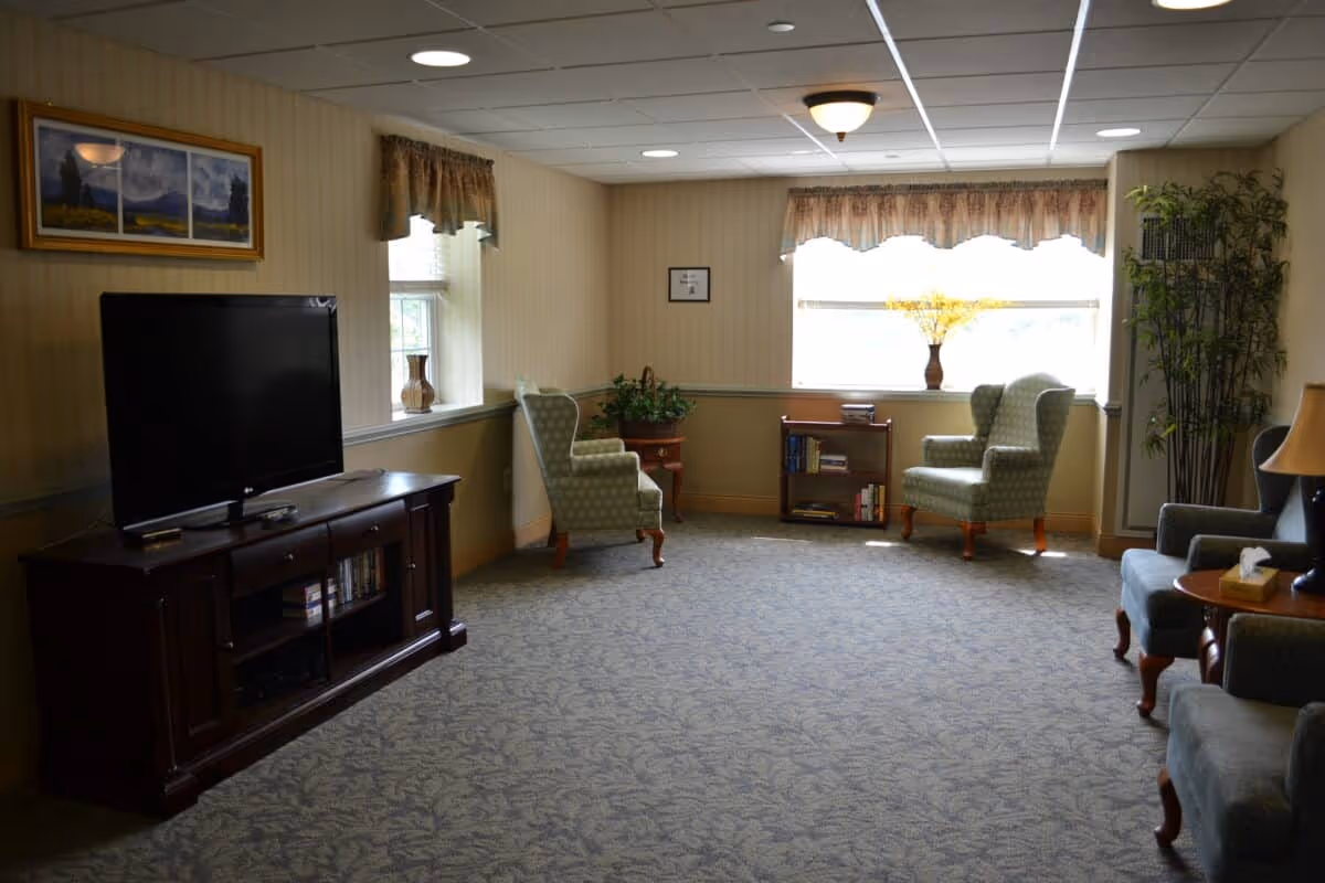 A cozy living room in Exton Senior Living with patterned carpet, two green upholstered armchairs near a window with floral valances, a wooden TV stand with a flat-screen television, a small bookshelf, a side table with a lamp, and decorative plants.