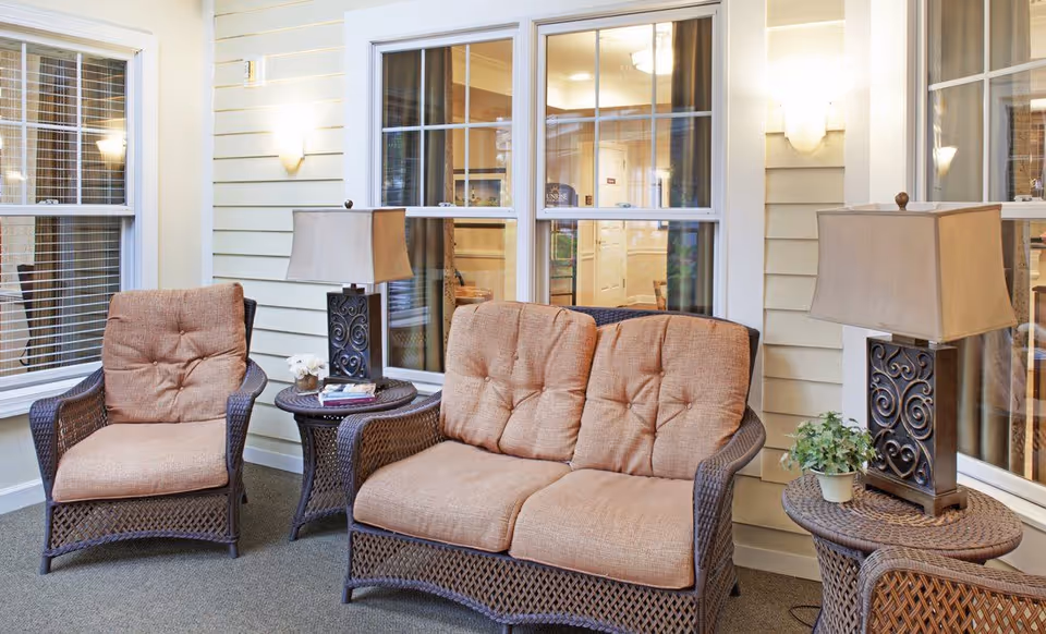Wicker loveseat and chairs with tan cushions and side tables and lamps in a bright indoor sitting area with windows.