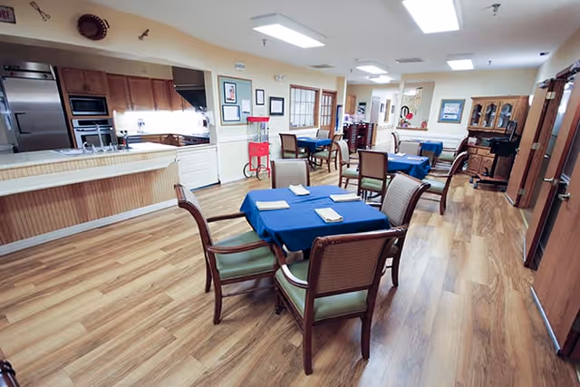 A spacious dining area with several tables covered in blue tablecloths, each set with napkins. The room features wooden flooring, wooden chairs with green cushions, and a kitchen area with stainless steel appliances and wooden cabinets. The walls are decorated with framed pictures and a popcorn machine is visible near the kitchen counter.