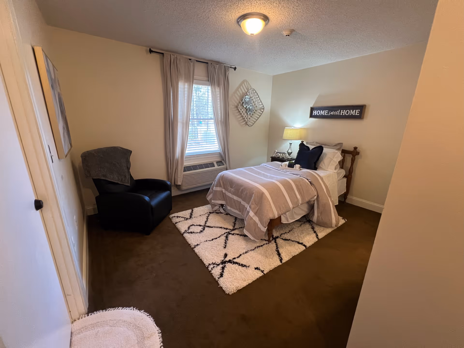A cozy bedroom in a senior living community featuring a single bed with striped bedding and multiple pillows, a bedside table with a lamp and decorative items, a comfortable black armchair with a blanket draped over it, a window with curtains, a wall decoration, and a 'Home Sweet Home' sign on the wall.