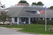 Single-story light-colored building with a covered entrance, American flag on a flagpole, driveway, and manicured lawn.
