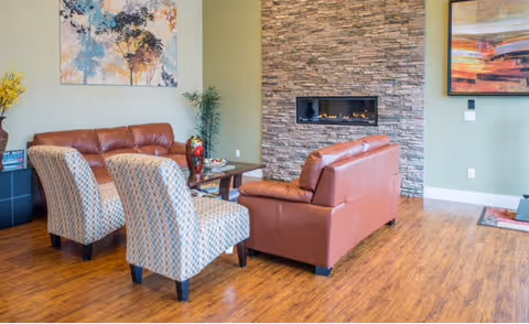 A cozy living room with two brown leather sofas and two patterned armchairs arranged around a wooden coffee table. The room features a stone fireplace built into a wall, wooden flooring, and light green walls adorned with colorful paintings. A small plant and decorative items are also visible.