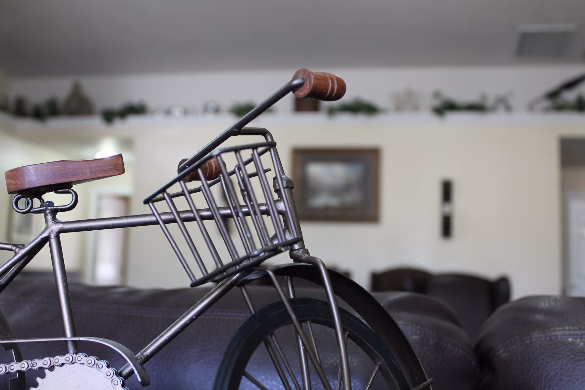 Close-up of a decorative metal bicycle with wooden seat and handle grips placed indoors, with a blurred background showing a living room area with couches, a framed picture on the wall, and some greenery on a shelf.