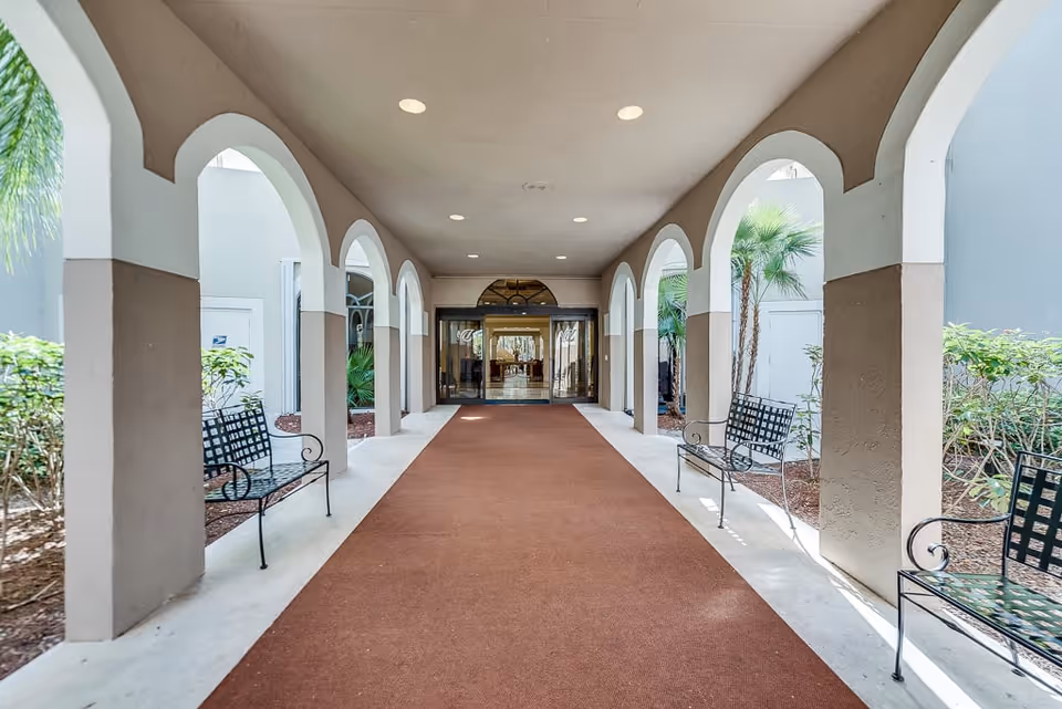 Covered walkway with arched openings on both sides, lined with black metal benches and plants outside the arches, leading to glass doors at the entrance of a building.