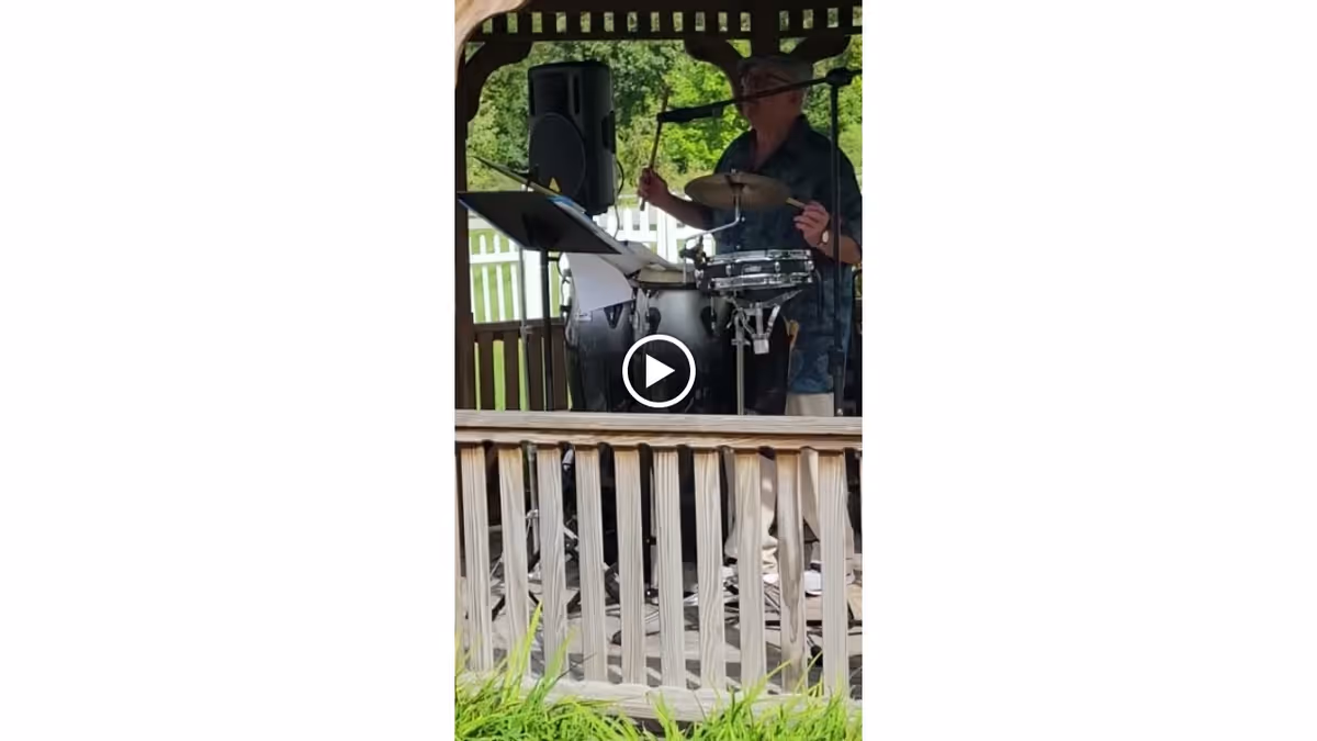 A man playing drums under a wooden gazebo with a white picket fence and green trees in the background.