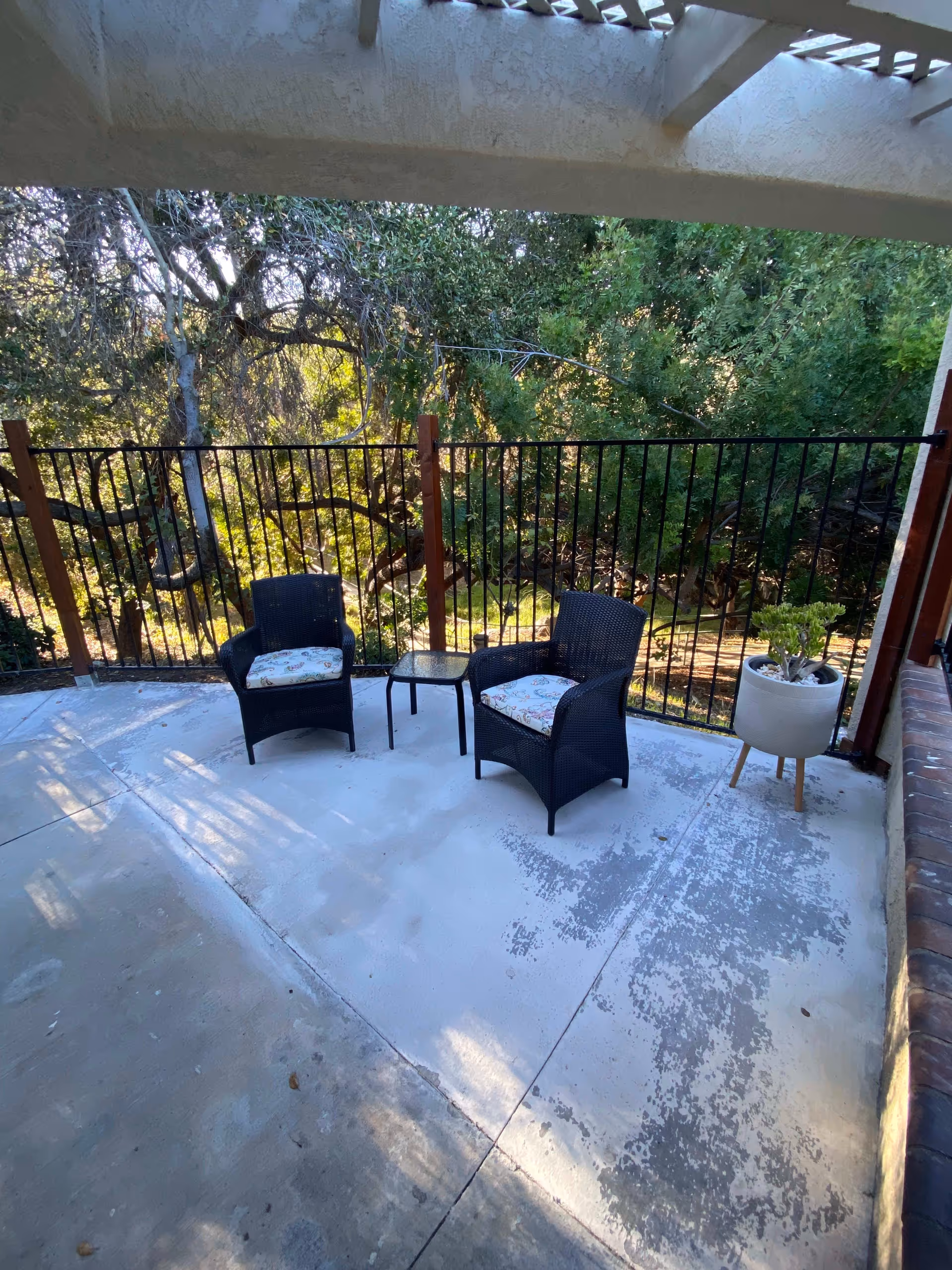 Outdoor patio area with two black wicker chairs with patterned cushions, a small glass-top table between them, and a potted plant on a stand. The patio is enclosed by a black metal fence and surrounded by trees and greenery.