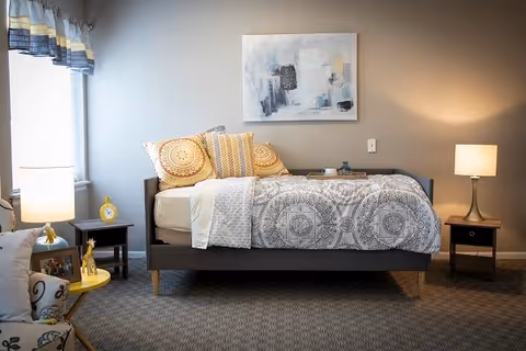A neatly made single bed with patterned bedding and decorative pillows in a softly lit bedroom. The bed is flanked by two nightstands, each with a lamp. A window with a valance is on the left side, and a piece of abstract art hangs on the wall above the bed. The room has a cozy and inviting atmosphere.