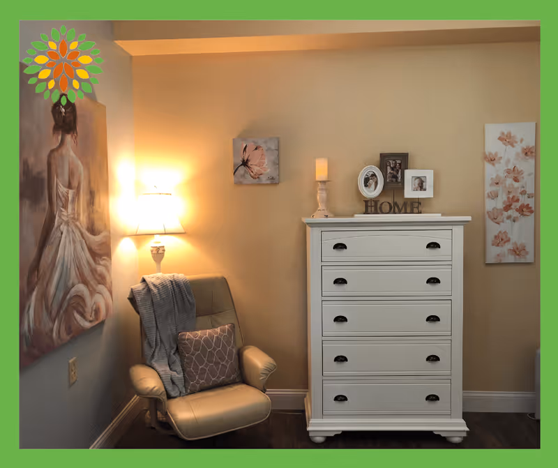 A cozy corner of a room featuring a beige armchair with a gray throw blanket and patterned pillow, a white chest of drawers with decorative items including framed photos, a candle, and a 'HOME' sign on top. The walls are adorned with three floral-themed paintings, and a wall lamp emits a warm light.