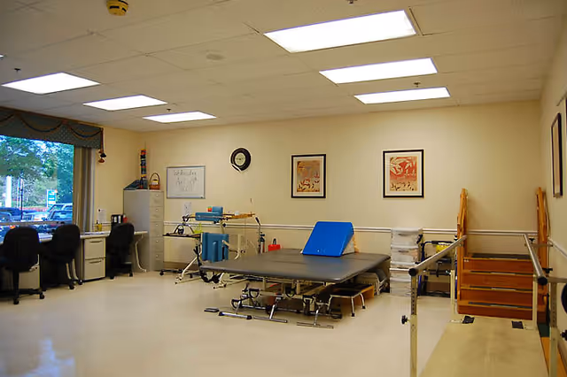 A physical therapy room with exercise equipment including parallel bars, a therapy table with a blue wedge cushion, a set of wooden stairs with handrails, two desks with chairs near a large window, and framed artwork on the walls.