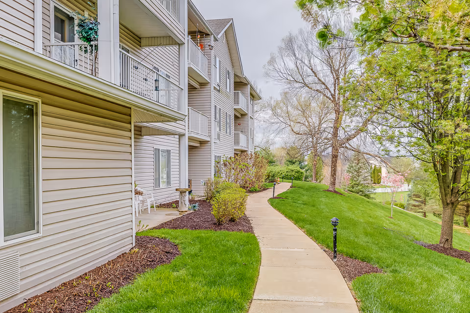 A paved walkway curves alongside a multi-story residential building with balconies. The area is landscaped with green grass, bushes, and trees, some of which are leafless, under a cloudy sky.