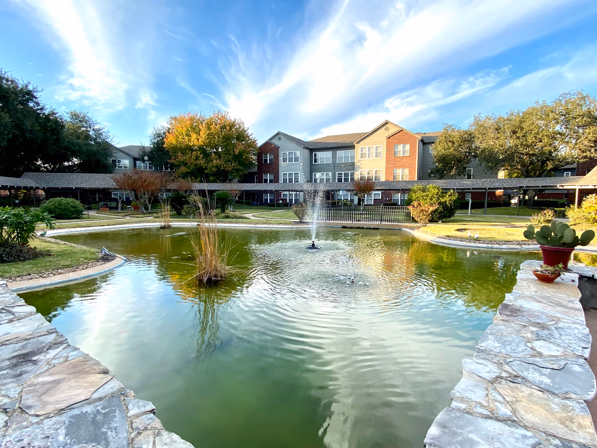 Outdoor view of a senior living facility with a large pond featuring fountains in the foreground, surrounded by stone edging and greenery. In the background, there is a three-story building with multiple windows and a covered walkway. Trees and bushes are scattered around the grassy area under a partly cloudy blue sky.
