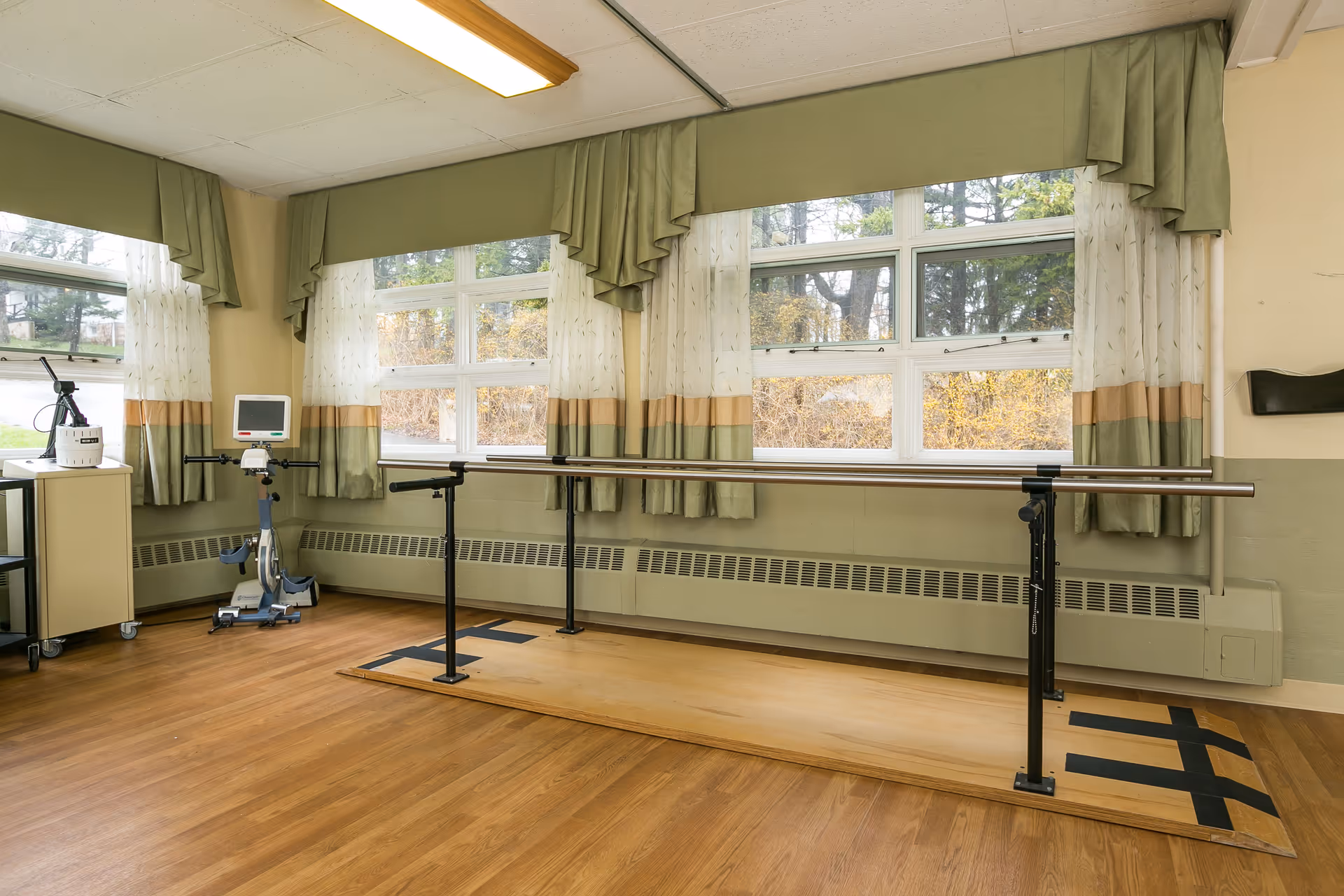 A bright rehabilitation room with parallel walking bars, an exercise bike, and large curtained windows.