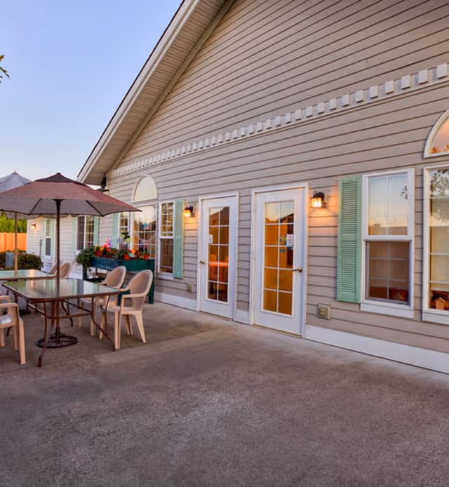 Outdoor patio area of a senior living facility with a large table, several plastic chairs, and a large umbrella. The building exterior features beige siding, green shutters, multiple windows, and glass doors with outdoor wall lights.