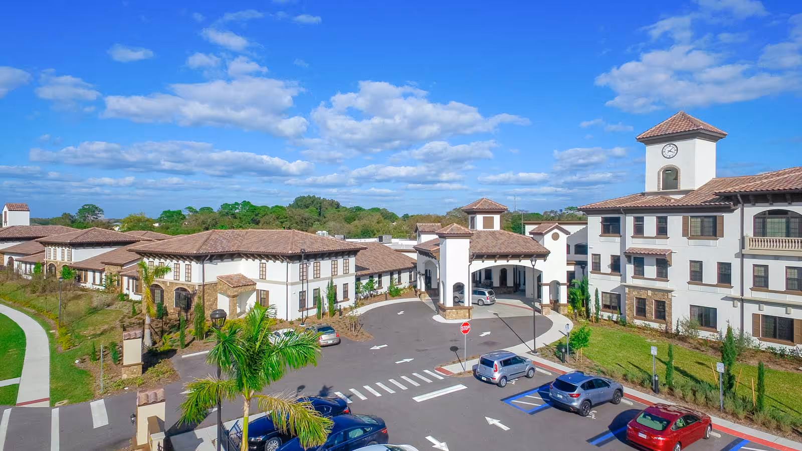 Exterior view of Tuscan Gardens of Venetia Bay Senior Living facility showing a large building with Mediterranean-style architecture, including terracotta roof tiles and a clock tower. The parking lot in front has several cars parked, and there are palm trees and landscaped greenery around the building under a partly cloudy blue sky.