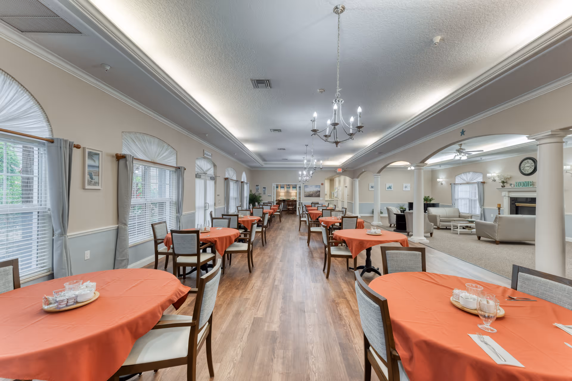 A spacious dining room with multiple round tables covered with orange tablecloths, each set with glasses, napkins, and condiments. The room has large windows with white blinds and light gray curtains, wooden flooring, and chandeliers hanging from the ceiling. To the right, there is a living area with sofas, a fireplace, and a clock on the wall.