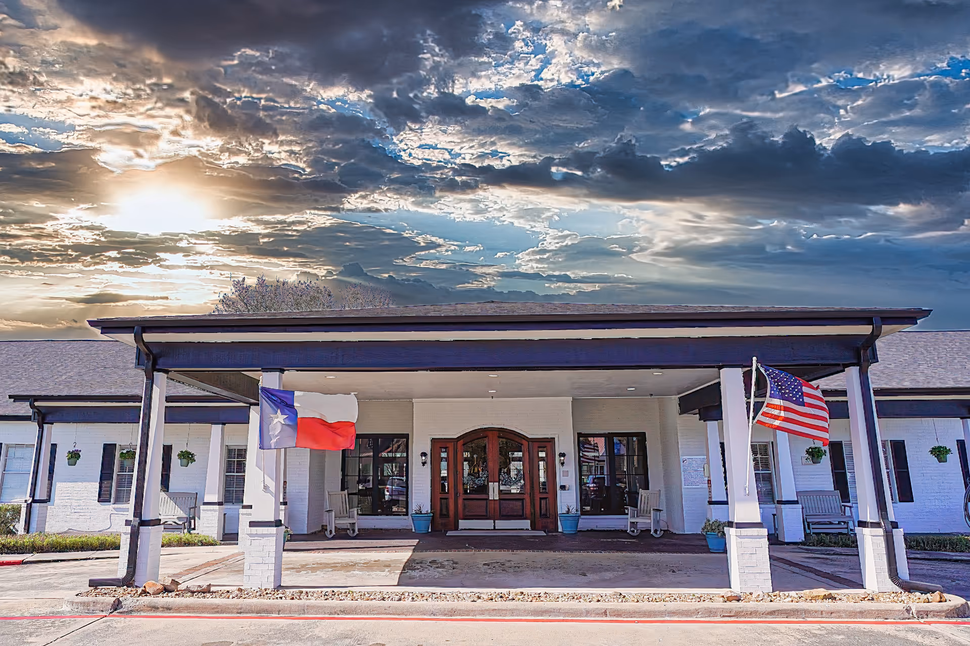Front entrance of a white senior living building with a covered porte-cochere and American and Texas flags under a dramatic cloudy sky.